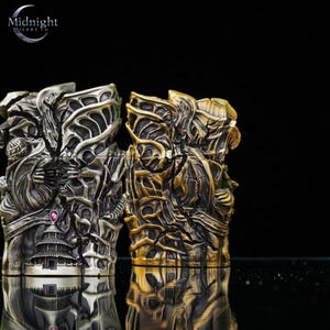 K&ouml;nnte beinhalten: Zwei kunstvoll gestaltete Metallfeuerzeuge, eines silberfarben und eines goldfarben. Das silberne Feuerzeug zeigt eine Pagode und einen rosa Edelstein. Beide Feuerzeuge haben detaillierte, strukturierte Oberfl&auml;chen mit einem k&uuml;nstlerischen Design. Das Logo von Midnight Trade Co. ist sichtbar.