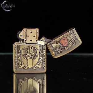 May include: A gold-colored metal lighter with an open lid. The lighter features intricate engraved designs, including a crest with two figures on horseback. The lid displays a shield with a red and orange emblem. The brand name "Midnight" is visible.