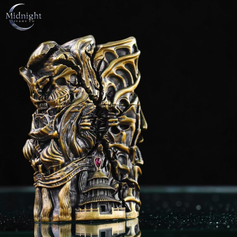 K&ouml;nnte beinhalten: Aufwendiges, bronzefarbenes Feuerzeug mit komplizierten Schnitzereien von Sch&auml;deln, Figuren und einer Pagode. Das Feuerzeug hat eine detaillierte, strukturierte Oberfl&auml;che und einen kleinen roten Edelstein. Das Logo von Midnight Trade Co. ist sichtbar.