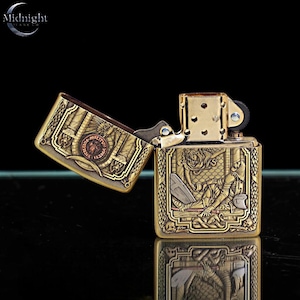 May include: A gold-colored metal lighter with intricate engraved designs. The lighter is open, revealing detailed artwork on both the lid and the body. The designs include figures, architectural elements, and text. The brand name "Midnight" is visible.
