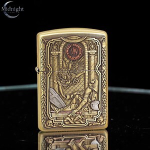 May include: A brass-colored lighter with an ornate engraved design. The design features a skeleton, an angel, and architectural elements. The lighter has the Midnight logo in the upper left corner. The lighter is rectangular and has a hinged lid.