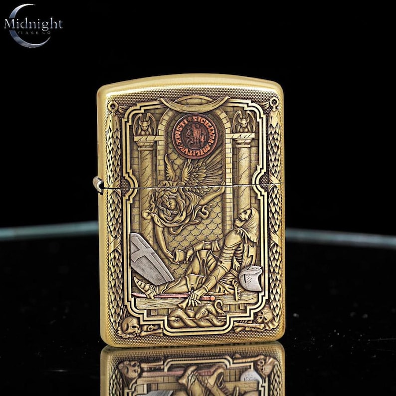 May include: A brass-colored lighter with an ornate engraved design. The design features a skeleton, an angel, and architectural elements. The lighter has the Midnight logo in the upper left corner. The lighter is rectangular and has a hinged lid.