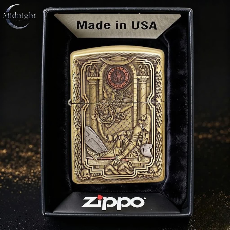 May include: A gold-toned Zippo lighter with an engraved design featuring a winged figure, a skeleton, and architectural elements. The lighter is presented in a black box, with the text "Made in USA" visible.