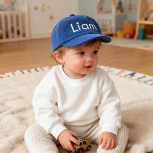 Puede incluir: Gorra de béisbol de pana azul con el nombre "Liam" bordado en blanco. La gorra la lleva un bebé que viste una sudadera blanca y pantalones beiges. El bebé sostiene un coche de juguete.