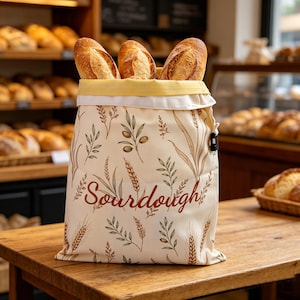 Puede incluir: Una bolsa de tela color crema con un estampado botánico de tallos de trigo y ramas de olivo, con la palabra Sourdough impresa en letra cursiva marrón. La bolsa contiene tres hogazas de pan crujiente y tiene un forro interior amarillo.