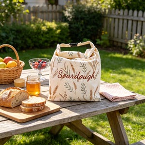 Puede incluir: Una bolsa de tela color crema para pan con cierre enrollable y hebilla de plástico negra. La bolsa presenta un patrón repetitivo de espigas de trigo y hojas en marrón y verde, con la palabra Sourdough impresa en cursiva marrón al frente.
