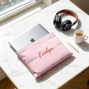 Può includere: Una custodia imbottita per laptop color rosa chiaro con il nome Evelyn ricamato in corsivo rosso, contenente un laptop argentato. Vicino ci sono cuffie nere, una tazza bianca riempita di caffè e una penna dorata.