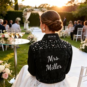 Puede incluir: Chaqueta vaquera negra adornada con perlas y el texto "Mrs. Miller" en escritura blanca. La fecha "05.04.26" está bordada en la parte superior de la espalda. La chaqueta se usa en una ceremonia de boda al aire libre.