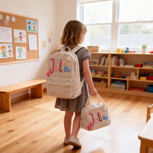 Puede incluir: Una mochila beige y una bolsa de almuerzo a juego, ambas personalizadas con el nombre "Julia" en bordado colorido. La mochila tiene un bolsillo frontal y adornos florales. La bolsa de almuerzo tiene un asa superior y un diseño floral similar.