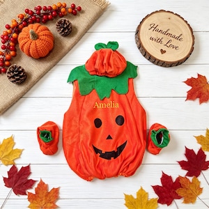 Puede incluir: Un pelele de bebé naranja con forma de calabaza, con gorro y patucos a juego. El pelele presenta una cara de calabaza negra y el nombre Amelia bordado en amarillo. El conjunto se muestra con hojas de otoño decorativas, piñas y bayas.