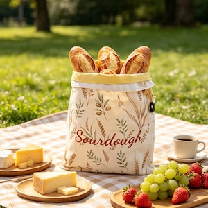 Può includere: Un sacchetto per il pane in tessuto color crema con una stampa botanica di steli di grano e rami d'ulivo. La parola Sourdough è stampata in corsivo rosso sul davanti. Il sacchetto ha una fodera interna gialla e contiene tre baguette dorate.