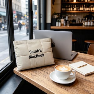 May include: A beige quilted laptop sleeve featuring the black embroidered text Sarah's MacBook, positioned next to a silver laptop, a white ceramic cup filled with latte art coffee, and a spiral-bound notebook with a gold pen on a wooden table.