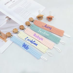 Puede incluir: Una colección de pinzas para chupetes de bebé en varios colores pastel, incluyendo azul, amarillo, rosa, verde y melocotón. Cada pinza tiene una textura similar a un gofre y presenta un nombre bordado en un color contrastante. También se ven cuentas de madera y chupetes.