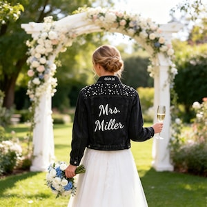 Puede incluir: Una chaqueta de mezclilla negra con adornos de perlas blancas en los hombros y la espalda. El texto "Mrs. Miller" está impreso en blanco en la parte posterior, con la fecha "05.04.26" bordada en el cuello. Se lleva sobre una falda blanca.