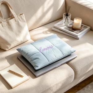Peut inclure: Une housse d'ordinateur portable matelassée bleu clair avec le prénom Emma brodé en lettres cursives violettes. La housse rectangulaire est fabriquée dans un tissu doux et rembourré, posée à plat pour offrir une protection à un ordinateur.