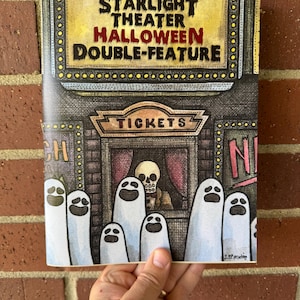 May include: A Halloween-themed book cover with the text "Starlight Theater Halloween Double-Feature." The illustration depicts a theater with ghosts and a skull at the ticket window. The cover is held against a brick wall.