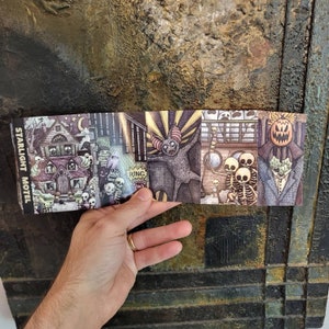 May include: A folded paper featuring a series of spooky illustrations. The images include a haunted house, skeletons, a bat-like creature, and a pumpkin-headed figure. The text "Starlight Motel" is visible on the left side of the paper.