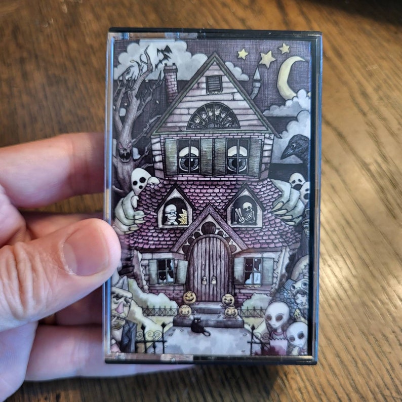 May include: A cassette tape with a spooky illustration of a haunted house. The artwork features ghosts, a black cat, pumpkins, and a full moon. The house has a pink roof and is surrounded by a spooky forest. The overall theme is Halloween.