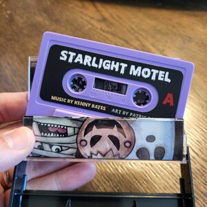 May include: A purple cassette tape labeled "STARLIGHT MOTEL" with black text. The cassette case has a colorful illustration of cartoon characters. The text "MUSIC BY KENNY BATES" and "ART BY PATRIC" are also visible.