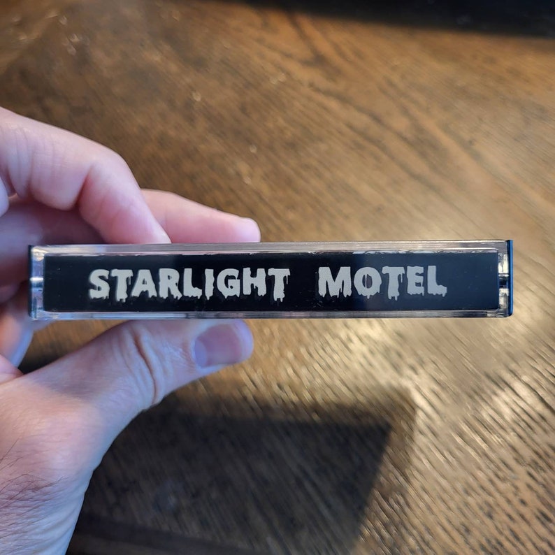 May include: A clear cassette tape with a black label. The label features the white text "STARLIGHT MOTEL" in a stylized font. The tape is held in a hand, with a wooden surface in the background.