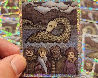 May include: A sticker featuring a cartoon illustration of a group of people standing in front of a large, spotted, serpentine creature in a body of water. The creature has a long neck and a small head with a friendly expression. The people are wearing colorful clothing and have a variety of hairstyles. The sticker has a glittery border.