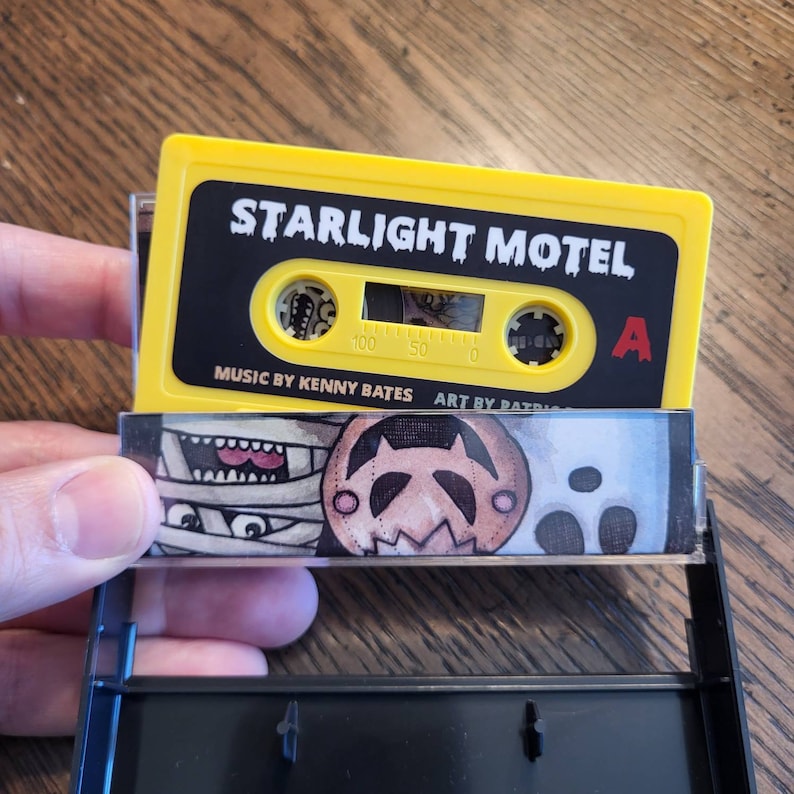 May include: A yellow cassette tape labeled "STARLIGHT MOTEL" with black text. The cassette case features a mummy, jack-o'-lantern, and ghost illustration. The text "MUSIC BY KENNY BATES" and "ART BY PATRICIA" are also visible.
