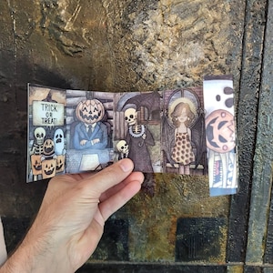 May include: A Halloween-themed paper art piece featuring a pumpkin-headed figure in a suit, skeletons, ghosts, and a winged woman. The artwork includes the text "Trick or Treat!" and is held against a textured background.