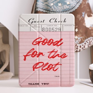 Puede incluir: Una funda para tableta rosa y blanca con las palabras "Good for the Plot" en escritura roja. La funda se asemeja a un recibo de cliente, con líneas y las palabras "Guest Check" en la parte superior. Cerca hay una pequeña taza marrón con una sustancia blanca similar a espuma.