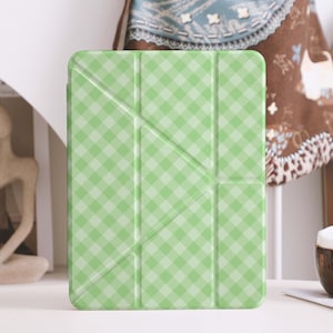 Op de afbeelding: Een lichtgroene tablethoes met een ruitpatroon. De hoes is opgevouwen en heeft een geometrisch ontwerp. Een bruine keramische beker met een lepel staat op een wit oppervlak. Een tijdschrift met het woord "MAGAZ" staat op de achtergrond.