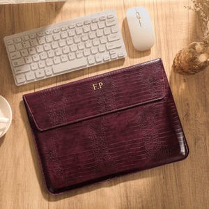 May include: A dark burgundy laptop sleeve with a textured crocodile skin pattern and the gold embossed initials F.P. on the front flap. The rectangular case features a fold-over closure and is positioned on a wooden surface near a keyboard.