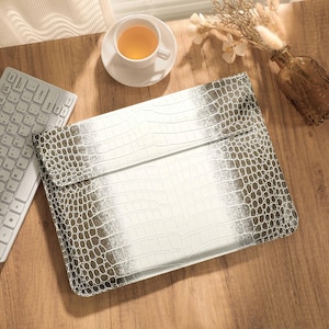 May include: A rectangular laptop sleeve featuring a metallic silver finish and a textured crocodile skin pattern. The sleeve has a fold-over flap closure and is positioned next to a white wireless computer keyboard on a wooden surface.