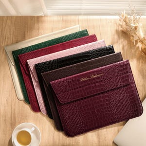 May include: A stack of six rectangular laptop sleeves featuring a textured crocodile skin pattern in cream, dark green, maroon, light pink, black, and dark brown. The front-most dark brown sleeve displays the gold embossed name Chloe Patterson.