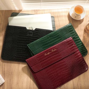 May include: Three rectangular laptop sleeves in black, dark green, and burgundy, each featuring a textured crocodile skin pattern. The burgundy sleeve is personalized with the gold-colored text Evelyn Porter embossed on the front flap.