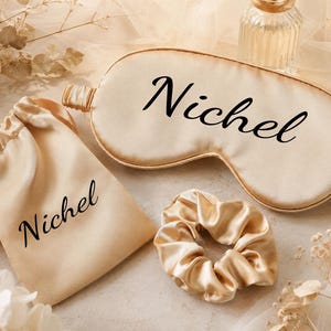 May include: A champagne-colored sleep mask, a matching drawstring pouch, and a scrunchie. The sleep mask and pouch both feature the name Nichel printed in a black cursive font. The items are made of a smooth, shiny fabric with an elastic strap.