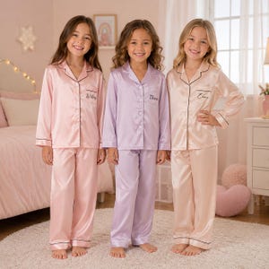 May include: Three sets of long-sleeved button-down pyjamas in satin fabric. The left set is light pink with the name Wiley, the centre set is lavender with the name Thea, and the right set is champagne with the name Emma. Each has black piping.