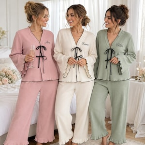 May include: Three sets of long-sleeved pyjama tops and wide-leg trousers in dusty rose, cream, and sage green. Each top features a V-neckline, black ribbon tie closure, lettuce-edge cuffs, and personalised embroidery reading Ava, Emma, or Chloe.