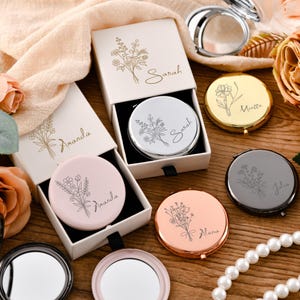 May include: A collection of round compact mirrors in silver, gold, rose gold, black, and matte pink finishes. Each mirror features a floral line art engraving and a name, including Sarah, Amanda, Miatta, Julia, and Alana, with matching gift boxes.