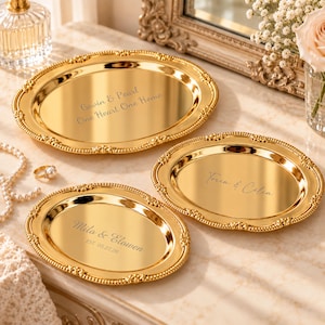 Puede incluir: Tres bandejas ovaladas de metal dorado con bordes de cuentas ornamentados. La bandeja más grande está grabada con "Gavin & Pearl One Heart One Home". La bandeja mediana dice "Torin & Celia". La más pequeña está grabada con "Milo & Elowen EST 05.27.26".