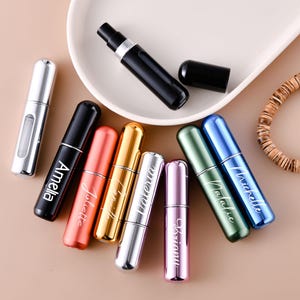 May include: A collection of small, cylindrical refillable perfume atomizer bottles in various metallic colors including silver, black, orange, gold, pink, green, and blue, some featuring white script names like Amelia, Juliette, and Michelle.