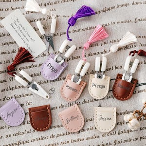 May include: A collection of small, white-handled scissors with silver blades, each in a colored leatherette pouch. The pouches are in various colors, with names like "Priya" and "Jessica" embossed on them. Tassels in various colors are also present.