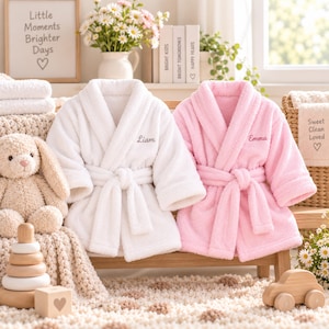May include: Two plush, long-sleeved bathrobes with tie belts, one in white with the name Liam embroidered on the chest and one in light pink with the name Emma embroidered on the chest. Both robes feature a shawl collar and soft, textured fabric.