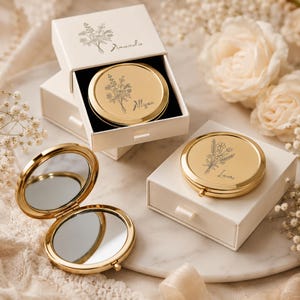 Puede incluir: Tres espejos de bolsillo circulares dorados con grabados de flores. Un espejo está abierto, mostrando dos superficies reflectantes. Dos espejos están cerrados, cada uno sobre una caja de regalo blanca deslizante con nombres y arte floral.