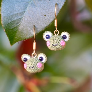 May include: A pair of dangle earrings featuring small, round, crocheted green frog heads. Each frog has white and dark blue eyes, pink stitched cheeks, and a black embroidered smile. The earrings hang from gold-toned metal fishhook wires.
