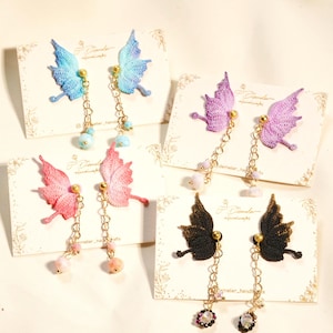 May include: Four pairs of butterfly wing earrings made of crocheted thread in blue, purple, pink, and black. Each pair features gold-tone metal chains with small dangling beads attached to the wings, displayed on individual white branded cards.