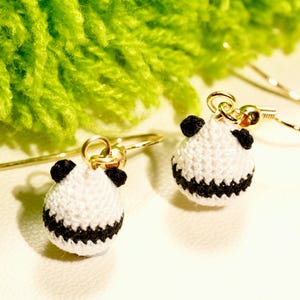 May include: A pair of dangle earrings featuring small, spherical, crocheted panda heads in white and black thread. Each panda charm is attached to a gold-toned metal fishhook ear wire, showcasing detailed stitching and tiny black ears.