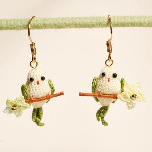 May include: A pair of dangle earrings featuring small, crocheted white birds with green wings and tails. Each bird is perched on a thin brown branch with a small, pale yellow flower attached. The earrings hang from gold-toned metal hooks.
