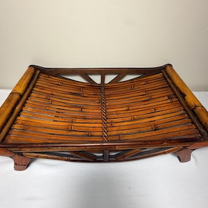 May include: A rectangular tray constructed from dark brown bamboo slats, featuring a curved, concave surface design. The tray is supported by four short, sturdy feet and includes a woven center seam detail along the length of the base.