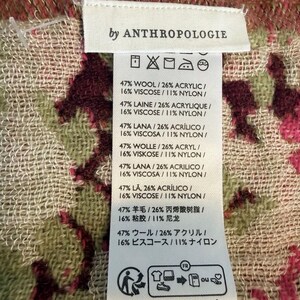 May include: A close-up of a tag from Anthropologie, featuring fabric composition details. The tag is white with black text, listing percentages of wool, acrylic, viscose, and nylon. The background shows a patterned fabric with pink and green floral designs.
