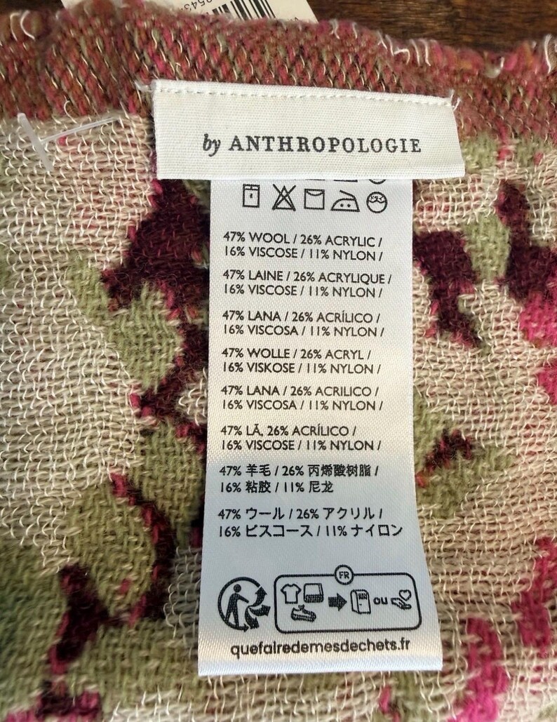 May include: A close-up of a tag from Anthropologie, featuring fabric composition details. The tag is white with black text, listing percentages of wool, acrylic, viscose, and nylon. The background shows a patterned fabric with pink and green floral designs.