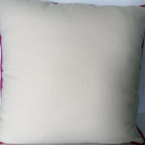 May include: Square decorative pillow with a cream-colored center and a vibrant magenta border. The pillow has a textured appearance, suggesting a natural fabric like linen or cotton. The design is simple and elegant, suitable for various home decor styles.